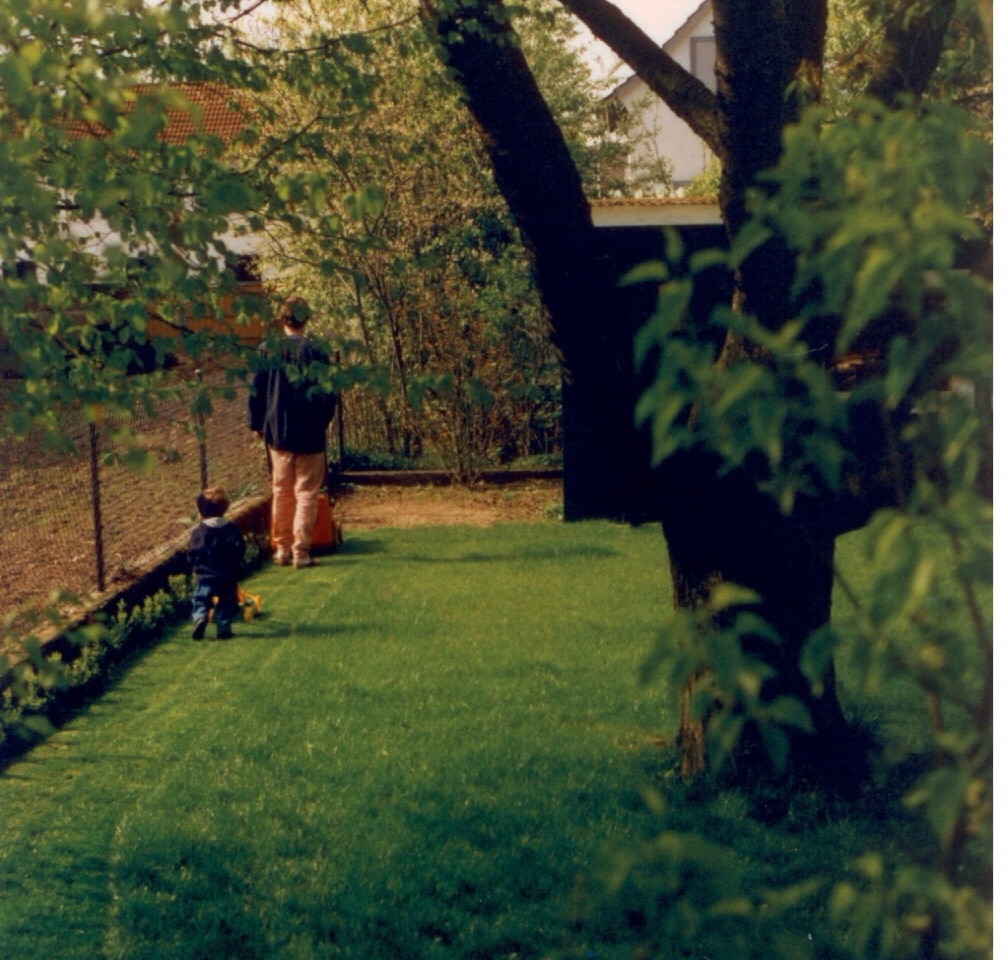 A toddler following an adult mowing a garden lawn